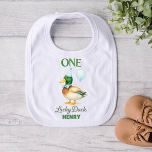 May include: A white baby bib featuring a mallard duck illustration wearing a blue party hat and bow tie, holding a light blue balloon. The text reads ONE, Lucky Duck, and HENRY in green and grey lettering centred on the fabric.