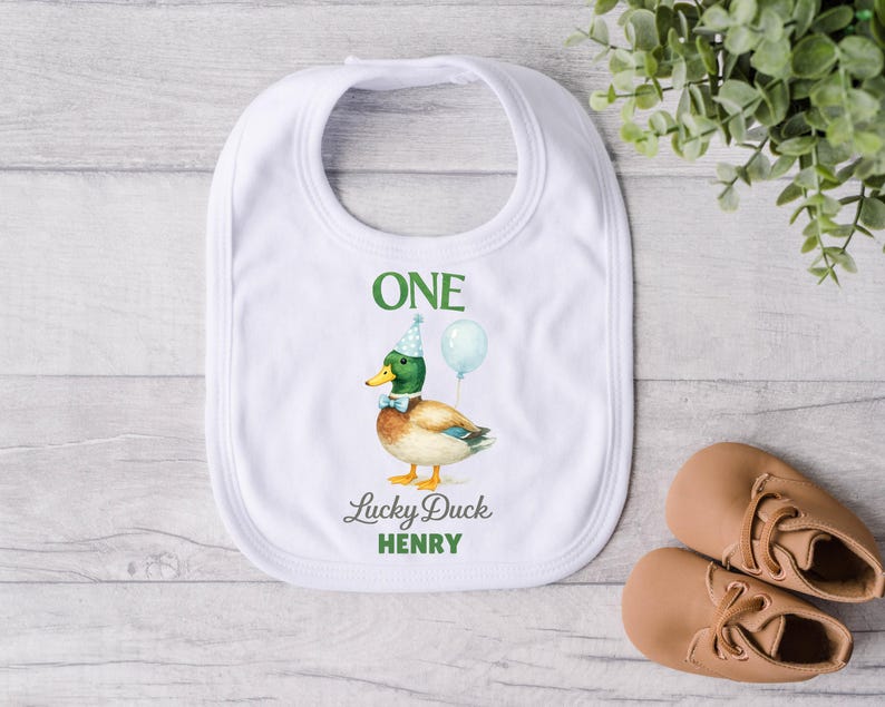 May include: A white baby bib featuring a mallard duck illustration wearing a blue party hat and bow tie, holding a light blue balloon. The text reads ONE, Lucky Duck, and HENRY in green and grey lettering centred on the fabric.