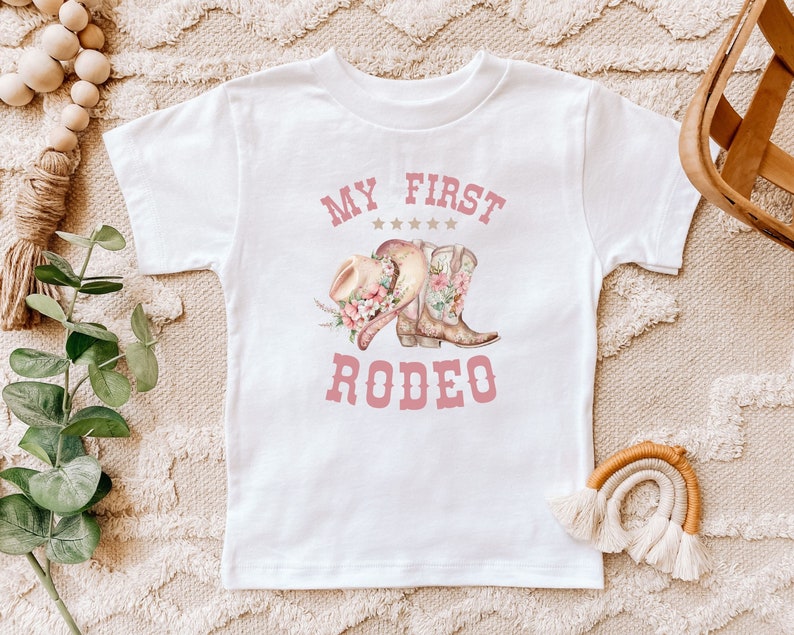 May include: White t-shirt with pink text that says "My First Rodeo" and a graphic of a cowboy hat and boots with floral accents.