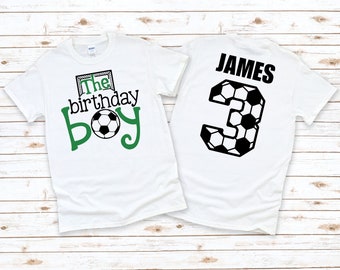 May include: Two white t-shirts with a soccer theme. The front shirt has green text that says "The birthday boy" with a soccer ball. The back shirt has the name "JAMES" and the number "3" made of soccer balls.