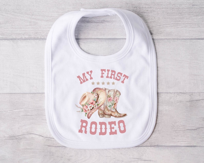 May include: White baby bib with pink text that reads "My First Rodeo" with a pink cowboy hat and boots with floral accents.