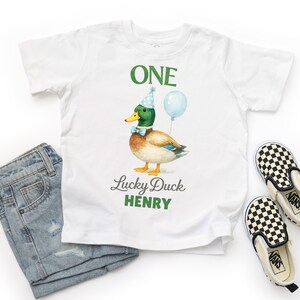 May include: A white t-shirt featuring a graphic of a mallard duck wearing a blue polka-dot party hat and a light blue bow tie, holding a light blue balloon. The text reads ONE, Lucky Duck, and HENRY in green and grey lettering.