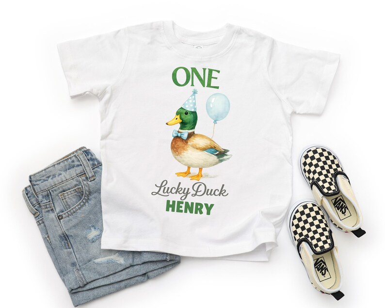 May include: A white t-shirt featuring a graphic of a mallard duck wearing a blue polka-dot party hat and a light blue bow tie, holding a light blue balloon. The text reads ONE, Lucky Duck, and HENRY in green and grey lettering.