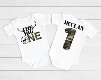 May include: Two white baby onesies. One onesie has a camouflage number one and the text "Declan". The other onesie has a deer head with antlers, a target, and the text "The Big One".