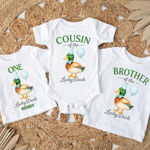 May include: Three white shirts and a bodysuit featuring a mallard duck wearing a blue party hat and bow tie with a light blue balloon. The text reads ONE Lucky Duck HENRY, COUSIN of the Lucky Duck, and BROTHER of the Lucky Duck in green.
