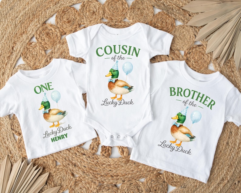 May include: Three white shirts and a bodysuit featuring a mallard duck wearing a blue party hat and bow tie with a light blue balloon. The text reads ONE Lucky Duck HENRY, COUSIN of the Lucky Duck, and BROTHER of the Lucky Duck in green.