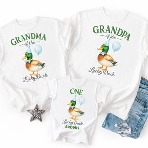 May include: Three white t-shirts featuring a mallard duck illustration wearing a blue party hat and bow tie with a light blue balloon. The shirts display the text Grandma of the Lucky Duck, Grandpa of the Lucky Duck, and One Lucky Duck Brooks.