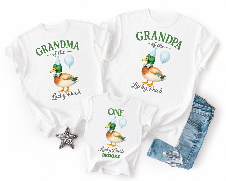 May include: Three white t-shirts featuring a mallard duck illustration wearing a blue party hat and bow tie with a light blue balloon. The shirts display the text Grandma of the Lucky Duck, Grandpa of the Lucky Duck, and One Lucky Duck Brooks.