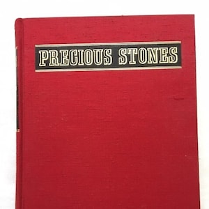 May include: A red hardcover book titled "PRECIOUS STONES" in gold lettering on a black banner. The book has a textured cover and a spine with black and gold accents. The book is closed and upright.