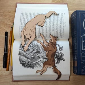 May include: Two paper cutouts of wolves, one brown and one white, are placed on an open book. The book is open to a page with an illustration of a man and a wolf.
