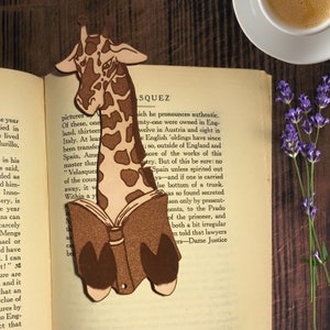 This cozy giraffe bookmark is made out of leather and is resting on the pages of an old book. Next to it on the right are some flowers and above that is a cup of coffee.