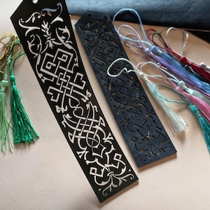 May include: Black laser-cut bookmarks with intricate white Celtic knot designs. The bookmarks have colorful tassels in shades of green, pink, blue, and white. The bookmarks are made of a dark material.