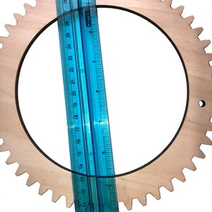 May include: A wooden gear with a diameter of approximately 14 inches. The gear is light brown and has a dark brown outline. A blue ruler is placed next to the gear for scale.