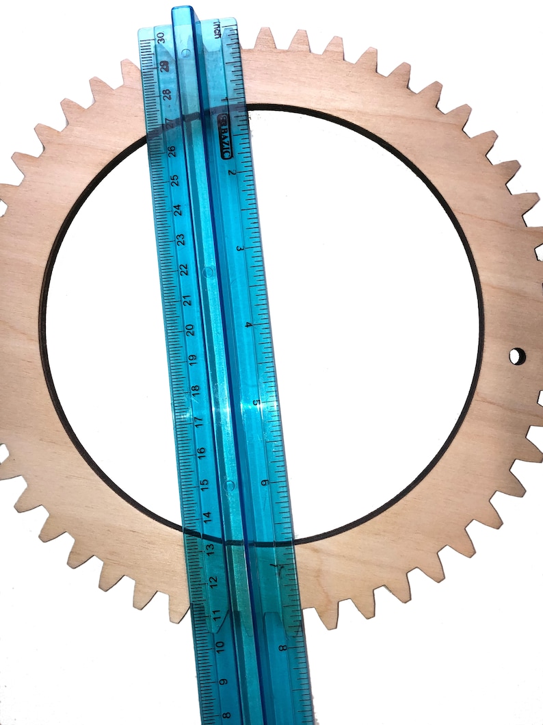 May include: A wooden gear with a diameter of approximately 14 inches. The gear is light brown and has a dark brown outline. A blue ruler is placed next to the gear for scale.