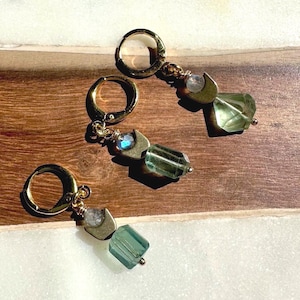 May include: Three gold-toned hoop earrings, each featuring a small iridescent bead and a crescent-shaped metal charm suspended above a faceted, translucent green rectangular gemstone. The earrings are arranged in a diagonal line on a surface.