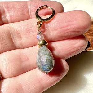 May include: A gold-toned hoop earring featuring a dangling teardrop-shaped labradorite gemstone. The design includes a small, clear faceted bead and a smooth gold-toned spacer bead positioned above the larger, iridescent gray labradorite stone.
