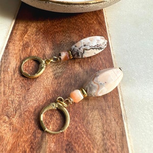 May include: A pair of gold hoop earrings with a pink and brown agate stone pendant. The earrings are on a wooden surface.