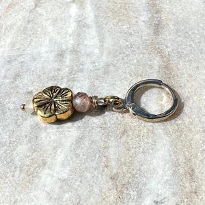 Puede incluir: Un solo pendiente dorado con un dije en forma de flor y pequeñas cuentas rosas. El pendiente tiene un cierre plateado. El dije de la flor tiene diseños detallados de pétalos. El pendiente está sobre una superficie clara y texturizada.