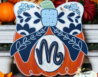 May include: A pumpkin-shaped wooden door hanger featuring a large dark blue bow with light blue floral accents. The center of the pumpkin displays a white circular cutout with a dark blue script letter M. The pumpkin has a light green stem.