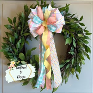 May include: A green leaf wreath with a large pastel bow. The bow features ribbons in pink, yellow, and blue with polka dots and plaid patterns. The text "Destined to Design" is on the lower left.