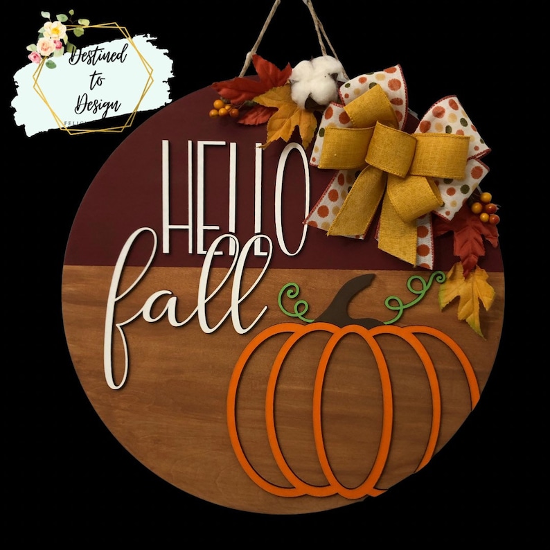May include: A round wooden door sign featuring a two-tone burgundy and brown background with the white text Hello fall. An orange pumpkin cutout, a large yellow burlap bow, artificial autumn leaves, and a white cotton boll are attached.