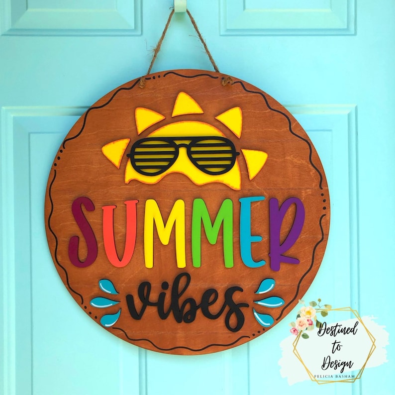 May include: A round wooden door hanger with a yellow sun wearing sunglasses and the text "SUMMER vibes" in rainbow colors.