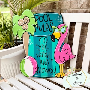 May include: A decorative wooden sign shaped like a pool float, featuring a teal background with the text "POOL RULES NO Y'ALL WATCH THIS! ALLOWED". It includes a bright pink flamingo wearing green sunglasses, a green palm leaf, and a beach ball.