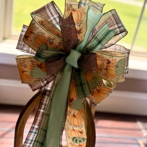 May include: A decorative bow crafted from multiple ribbons featuring a fall-themed pattern of orange pumpkins, brown leaves, and acorns, interspersed with solid sage green and brown burlap ribbons, along with brown and green plaid patterned ribbons.