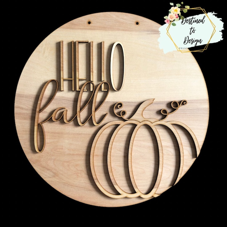 May include: A circular wooden wall sign featuring the text "HELLO fall" in a combination of block and script lettering. A stylised pumpkin outline is positioned below the text. The sign has two small holes at the top for hanging purposes.