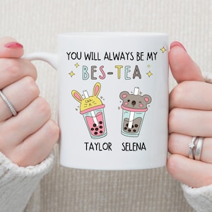Puede incluir: Taza de cerámica blanca con un contorno negro y el texto "You will always be my bes-tea" con dos tazas de té de burbujas de dibujos animados con los nombres "Taylor" y "Selena" debajo.