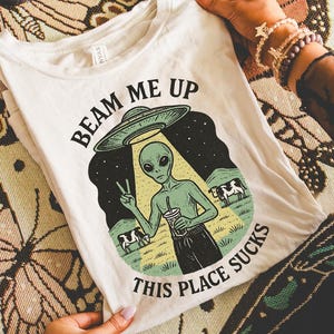 May include: Off-white t-shirt with the text "BEAM ME UP THIS PLACE SUCKS" and a graphic of an alien giving a peace sign. The alien is holding a drink, with cows in the background. The shirt is on a patterned surface.