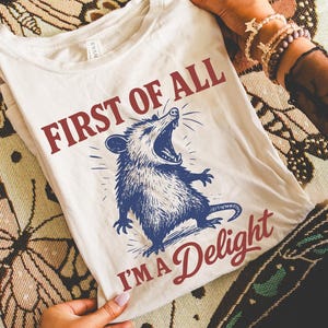 May include: Cream-colored t-shirt with the text "FIRST OF ALL I'M A Delight" and a blue illustration of an opossum with its mouth open. The shirt is displayed on a patterned surface with candles.