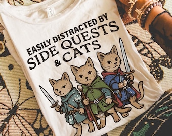 May include: Cream-colored t-shirt with the text "EASILY DISTRACTED BY SIDE QUESTS & CATS" and a graphic of three cartoon cats dressed as fantasy characters, each holding a sword or staff. The shirt is being held up, with candles and a patterned cloth in the background.