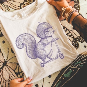 Puede incluir: Camiseta color crema con una ilustración morada de una ardilla con casco montada en un scooter. La camiseta gráfica es sostenida por una persona, con velas y una manta estampada en el fondo.