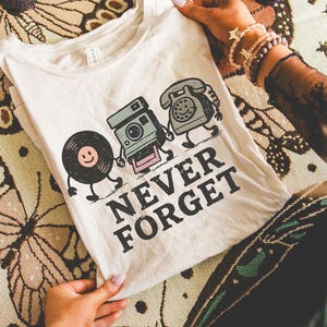 May include: Cream-colored t-shirt featuring a graphic of a smiling vinyl record, camera, and telephone walking together. The words "NEVER FORGET" are printed below the characters in black. The shirt is held up against a patterned background.