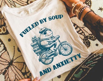 May include: Cream-coloured t-shirt with the text "Fueled by Soup and Anxietty". The shirt features a blue illustration of a frog riding a bicycle with a basket. The shirt is held up by a person.