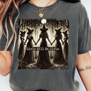 May include: Dark gray t-shirt featuring a graphic of five witches holding hands in a forest, with the text "Girls Will Be Girls." The shirt is part of the Pepper T-Shirt collection.