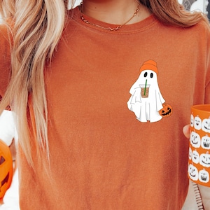 May include: Rust-coloured t-shirt with a ghost wearing an orange beanie, holding a drink and a pumpkin. A pumpkin-shaped candy holder and a mug with pumpkin faces are also visible. The text "YAM" is in a white box.