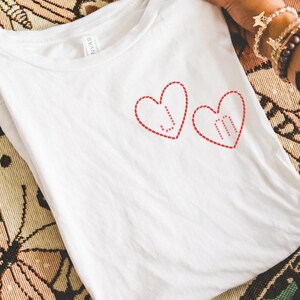 Peut inclure: Un t-shirt blanc avec deux motifs de cœur rouges, chacun contenant des initiales. Le t-shirt est plié et tenu par des mains, sur un fond à motifs avec des bougies. Le design est simple et romantique.