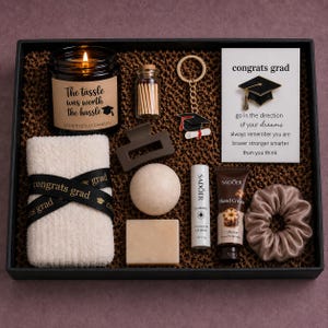 May include: A graduation gift set in a black box, featuring a candle, matches, a keychain, a towel with a ribbon, a hair clip, a bath bomb, a bar of soap, lip balm, hand cream, and a scrunchie. The set includes a card that says "congrats grad".