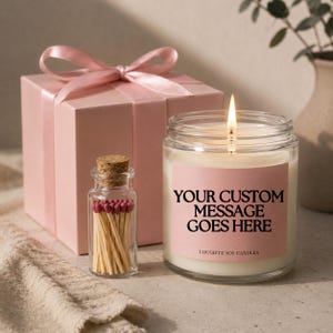 May include: A lit soy candle in a glass jar with a pink label that reads "YOUR CUSTOM MESSAGE GOES HERE". A small glass bottle of matches and a pink gift box with a ribbon are also present.