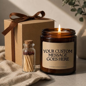 Peut inclure: Une bougie de soja allumée dans un bocal en verre brun avec une étiquette indiquant "YOUR CUSTOM MESSAGE GOES HERE". Une petite bouteille en verre d'allumettes et une boîte cadeau marron avec un ruban sont également présentes.