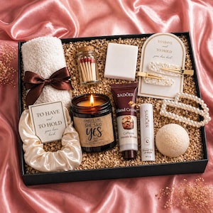May include: A black gift box filled with self-care products. It contains a fluffy white towel with a brown ribbon, a candle, hair clips, lip balm, hand cream, and a bath sponge. The candle says "Smells Like She Said Yes".