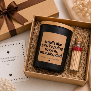 May include: A gift box containing a black candle with a wooden lid and a label reading 'smells like you're going to be an amazing dad', a small glass bottle filled with matches with red tips, and a white card with a heart border and text.