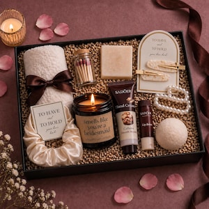 May include: A gift box containing self-care products. It features a candle, matches, soap, hair accessories, lip balm, hand cream, a towel, and a scrunchie. The candle has the text "smells like you're a bridesmaid".