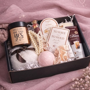 Puede incluir: Caja de regalo con una vela que dice "Smells Like She Said Yes", un collar de perlas, una bomba de baño y un tubo de crema de cafeína. La caja está sobre una tela rosa. Se ve la marca "Unicorntrendz".