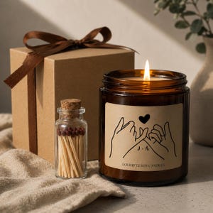 May include: A lit soy candle in a brown glass jar with a label featuring clasped hands and a heart. A small glass bottle of matches and a brown gift box with a ribbon are also present.