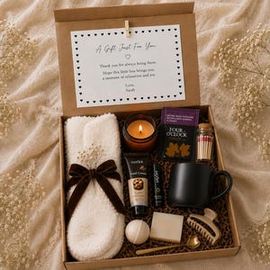 May include: A gift box containing a white fuzzy scarf with a brown velvet bow, a lit candle in a brown glass jar, a box of Four O'Clock tea, a small glass bottle of matches, a black mug, hand cream, lip balm, a bath bomb, soap, and a hair clip.