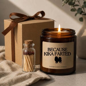 Peut inclure: Une bougie en verre brun allumée avec le texte "BECAUSE KIKA FARTED" et une silhouette de chien. Une petite bouteille d'allumettes et une boîte cadeau marron avec un ruban sont également présentes.