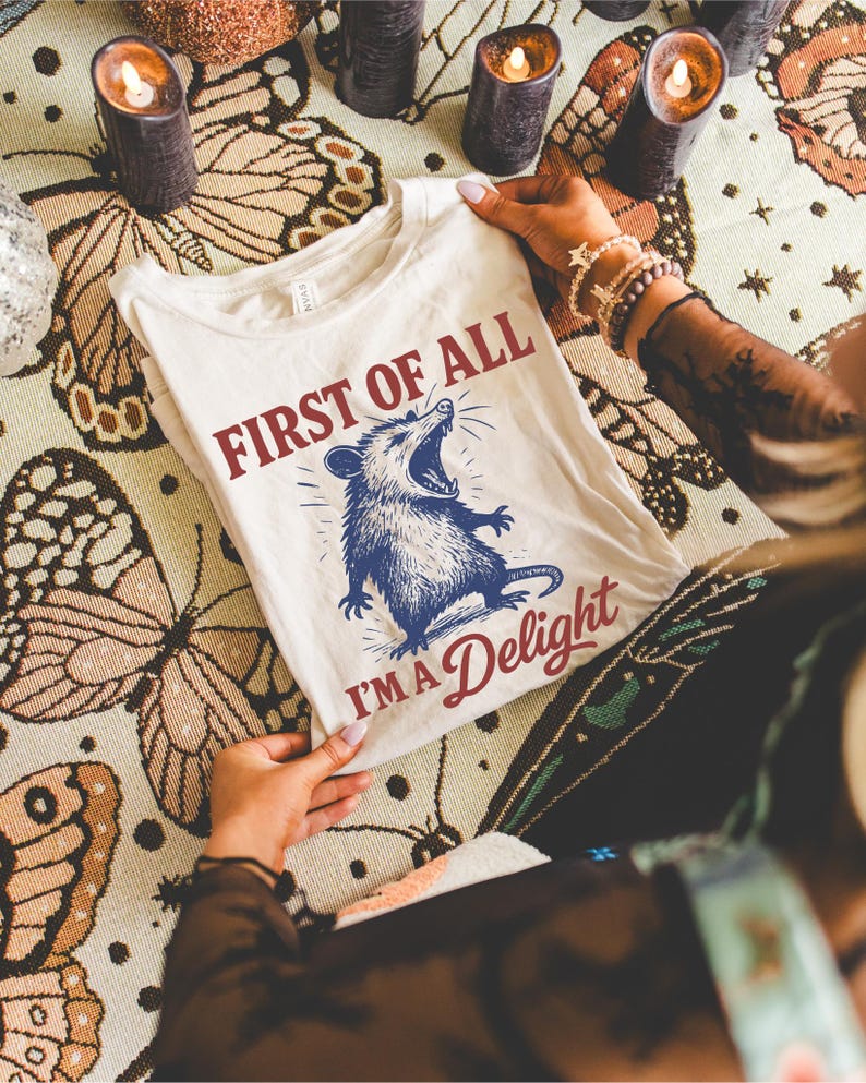 May include: Cream-colored t-shirt with the text "FIRST OF ALL I'M A Delight" and a blue illustration of an opossum with its mouth open. The shirt is displayed on a patterned surface with candles.